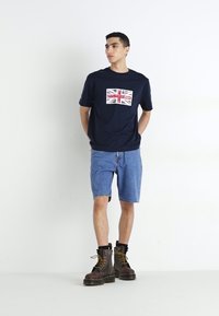 Jūras spēku t-krekls ar Union Jack grafiku, saskaņots ar gaiši zilām džinsu šortiem. Modelis valkā brūnas zeķes botas. Vienkārša baltā fona.