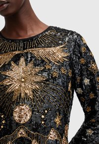 Top negru cu paiete, decorat cu embellismente aurii într-un design celestial, cu stele și motive florale ce împodobesc materialul. Mâneci lungi, decolteu rotund.