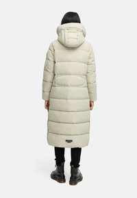 Abrigo de plumas largo en un tono beige claro, con capucha, que presenta segmentos acolchados horizontales y un parche de logo en el dobladillo. Combinado con pantalones oscuros y botas.