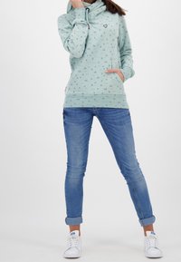 Sudadera verde menta con cuello de cowl, que presenta un pequeño diseño de corazón y un bolsillo tipo canguro, combinada con jeans ajustados azules y zapatillas blancas.