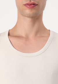Beige ribbed tank top with a wide crew neck, made of soft fabric, featuring a smooth texture and neat stitching around the neckline.