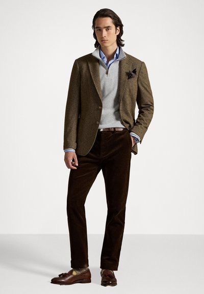 Άνδρας με καφέ σακάκι, ανοιχτό γκρι ζακέτα quarter-zip, μπλε πουκάμισο με γιακά, σκούρες καφέ παντελόνες και καφέ loafers με φούντες, που στέκεται μπροστά σε απλό φόντο.