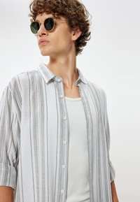 Camisa ligera de cuadros en blanco y gris, usada sobre una camiseta sin mangas blanca, complementada con gafas de sol redondas con lentes oscuros.