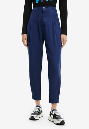 Persoon draagt high-waist, losse donkerblauwe broek met opgerolde pijpen, een zwarte top met lange mouwen en zwart-witte sneakers met dikke zolen.