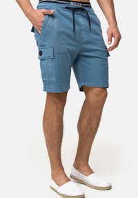Shorts cargo bleu clair en coton, avec des poches latérales, une taille à cordon de serrage et un ourlet cousu. Portés avec des chaussures blanches décontractées à enfiler.