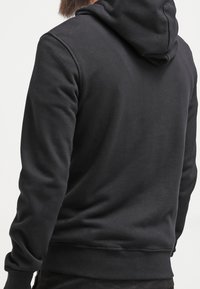 Sweat à capuche noir avec une texture douce, doté d'une capuche et de poignets côtelés. Design minimaliste, sans logos ni motifs visibles, coupe simple.