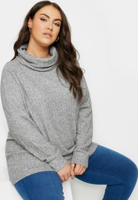 Maglione grigio a collo alto con una texture a coste e maniche lunghe, abbinato a jeans blu. Il tessuto sembra morbido e accogliente.