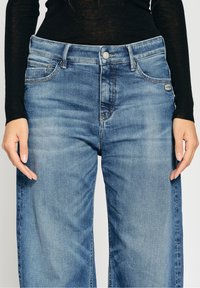 Sinisen farkkukankaan suorat lahkeet, viisi taskua, vaaleaa kulutusta ja hopeinen nappisulku. Vyötäröllä oleva etiketti esittää brändin logoa.