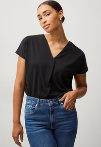 Sort kortærmet bluse med v-hals, der har blødt stof og en sammenkrøllet detalje ved taljen, parret med højtaljede blå jeans.