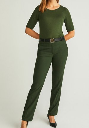Woman wearing dark green fitted half-sleeve top and matching trousers with black belt and black pointed high heels, standing against plain background.