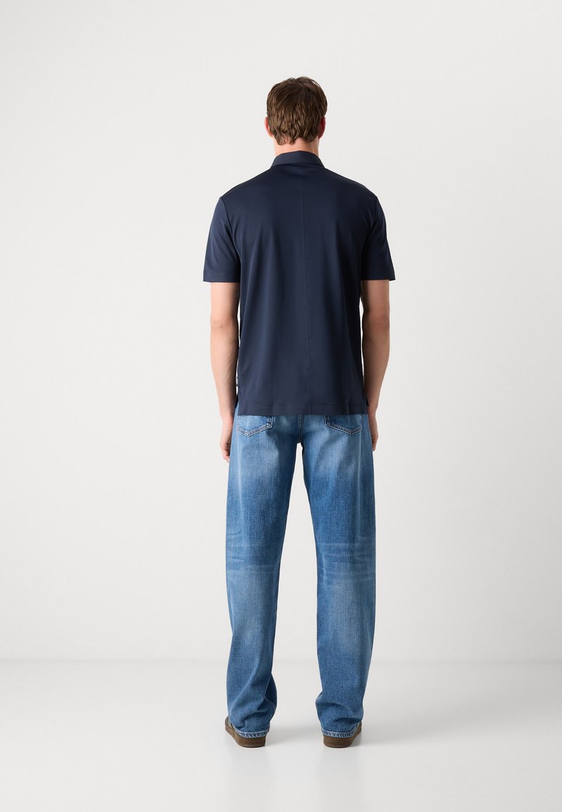 Polo shirt bleu marine avec un léger éclat, manches courtes et ourlet droit, associé à un jean en denim bleu clair, ample, présentant des zones de décoloration.