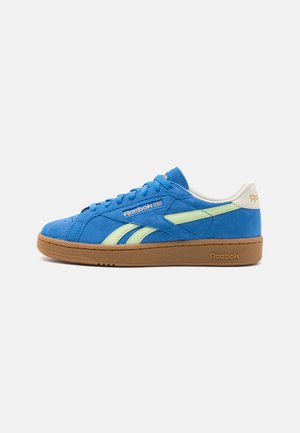 Zapatillas deportivas bajas de ante azul con detalles verdes, suela de goma y la marca Reebok en el lateral y el talón. Cintas Planas y cuello acolchado.