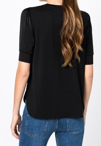 Svart kortärmad topp med rundad halsringning, veckade axlar och något böjd nederkant, matchad med blå jeans. Slät textur.