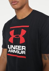 Camiseta de algodón negra con un atrevido logo rojo de Under Armour y texto blanco. Cuello redondo, mangas cortas y corte regular con una textura suave.