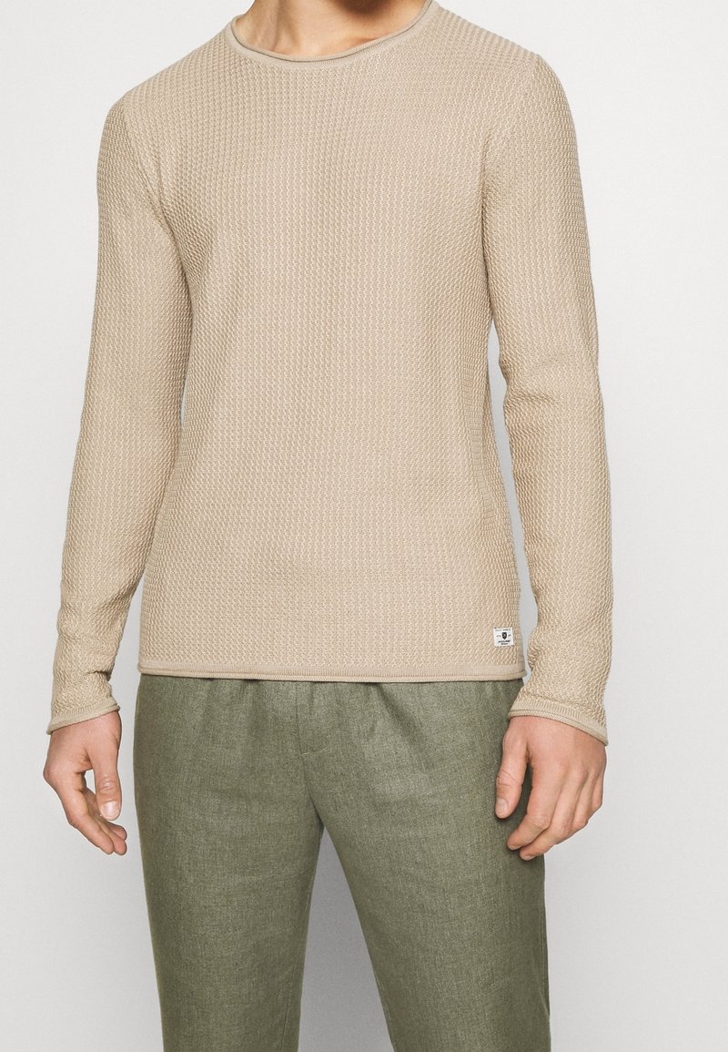 Pull beige clair à manches longues avec un design tricoté texturé et une petite étiquette logo près de l'ourlet. Associé à un pantalon vert olive.