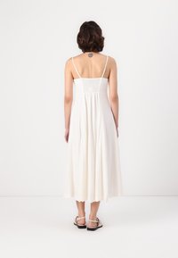 Abito lungo bianco con spalline sottili, corpetto smock e gonna svasata. Sandali con decorazioni e suole nere visibili sul fondo.