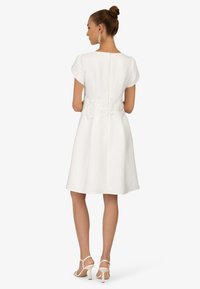 Robe blanche à manches courtes, avec des détails en dentelle à la taille et une fermeture éclair dissimulée dans le dos. Associée à des talons beiges à lanières.