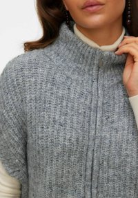 Kvinna som bär en tjock grå stickad tröja med dragkedja, en krämfärgad polo-tröja under och hängande mörka pärlörhängen.