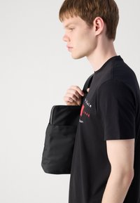 Zwarte stoffen tote bag met ritssluiting, rechthoekige vorm en eenvoudig design, vastgehouden door een persoon in een zwart t-shirt met rode accenten.