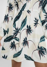 Blumenkleid aus leichter Stoffqualität mit grünen und pinken botanischen Prints auf cremefarbenem Hintergrund; elastischer Taillenbund und lange Ärmel.