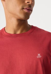 Camiseta de algodón roja con cuello redondo, que presenta un pequeño logotipo blanco "M O’P" cerca del área del pecho. Textura suave.