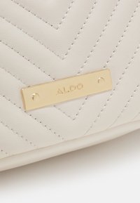 Krémově texturovaná kožená kabelka se vzorem chevron, na boku s kovovou zlatou značkou s raženým nápisem "ALDO".