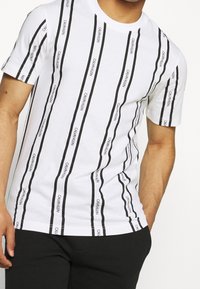Bílý krátký rukáv tričko s vertikálními černými pruhy a textem "CALVIN KLEIN". Měkká tkanina, kulatý výstřih, standardní střih.