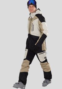 Lyžařský oblek s modulárním designem v béžové a černé barvě, se zipovými kapsami a helmou, doplněný brýlemi a lyžařskými botami.