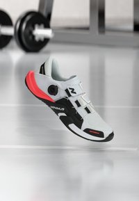 Light gray athletic shoe with a black and neon pink sole, textured upper, adjustable strap, and a circular fastening mechanism.