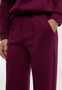 Pantaloni della tuta color bordeaux con una texture morbida, vita elasticizzata, tasche laterali e cuciture a contrasto lungo le giunture.