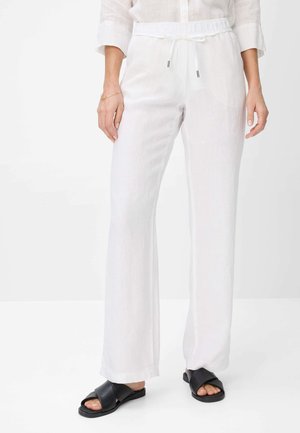 Vrouw draagt witte linnen broek met trekkoord en zwarte slides met gekruiste bandjes, staand tegen een effen witte achtergrond.