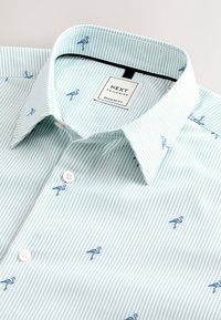 Camicia a maniche corte verde chiaro a righe con fenicotteri blu ricamati, colletto classico, pattina frontale con bottoni bianchi e etichetta del marchio.