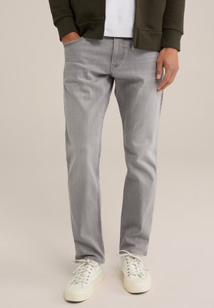 Man draagt lichtgrijze spijkerbroek, witte sneakers, wit shirt en donkergroene jas met rits, staat met één hand in de zak.