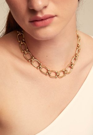 Collar de cadena de oro con eslabones ovalados y acentos redondeados, descansando sobre la piel. Textura suave, acabado brillante y elementos de diseño prominentes.
