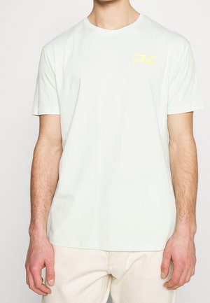 Homme portant un T-shirt à manches courtes de couleur unie claire avec un petit motif jaune sur la partie supérieure gauche de la poitrine et un pantalon clair.