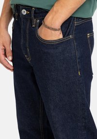 Tmavomodré denimové džínsy s žltým prešívaním, prednou kapsou a klasickým dizajnom s bežným pásom a zapínaním na gombík.