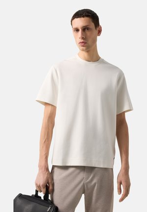 Ung mand iført en enkel hvid oversized T-shirt og beige bukser, der holder en lille sort lædertaske i venstre hånd.