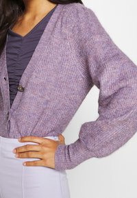 Ljuslila stickad cardigan med V-ringning och knappar framtill. Har texturerad tyg och åtsittande ärmar. Bärs över en mörk topp.