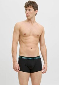 Ein oben ohne Mann trägt schwarze Boxer-Shorts von Jack & Jones mit einem blauen Logo-Bund und schaut nach links vor einem schlichten weißen Hintergrund.