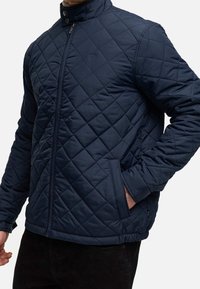 Homme portant une veste matelassée bleu marine avec fermeture éclair frontale et poches latérales, main dans la poche, sur un fond clair uni.
