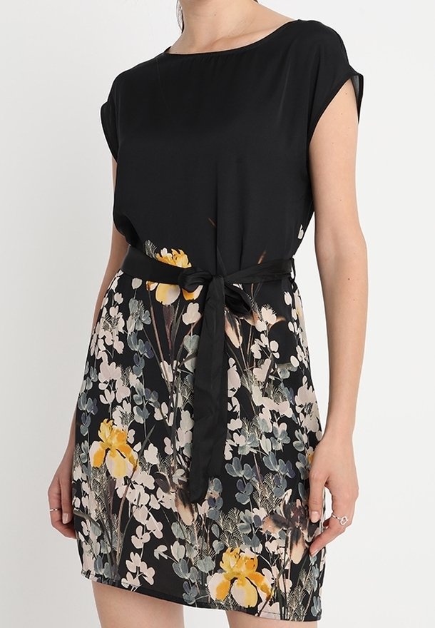 Mujer vestida con un vestido negro de mangas cortas, falda floral con flores amarillas y blancas, y un cinturón negro atado en la cintura.