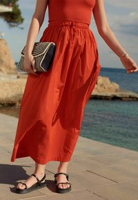 Falda maxi roja y fluida con bolsillos laterales, combinada con sandalias negras con tiras y un bolso de mano estampado que presenta rayas en azul marino y blanco.