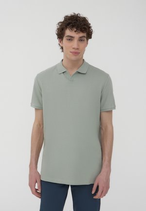 Polo shirt di colore verde chiaro, maniche corte, texture a coste, colletto classico, vestibilità rilassata. Indossato con pantaloni blu scuro.