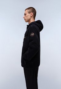 Black fleece jacket with a hood, featuring a zip pocket and textured fabric. Includes a round logo patch on the left sleeve.