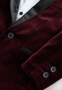 Satinblazer i velour burgunder med sorte satinlapeller, sorte knapper foran og ved ærmerne, samt en hvid skjorte under.