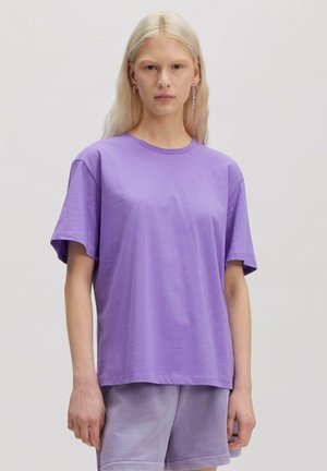 Persoon met lang blond haar die een effen paarse t-shirt en bijpassende paarse korte broek draagt tegen een effen achtergrond.