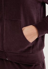Bordeaux kleurige zip-up hoodie met een voorzak, ribbelmanchetten en zachte, gestructureerde stof, gedeeltelijk getoond met een hand in de zak.