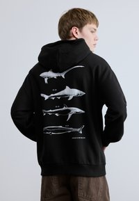 Μαύρο φούτερ με κουκούλα με ένα λευκό γραφικό που απεικονίζει τέσσερις καρχαρίες στην πλάτη, με ετικέτες και κείμενο που περιγράφουν την εξέλιξή τους.