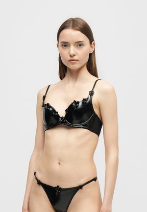 Jeune femme portant un soutien-gorge noir brillant et une culotte taille basse assortie avec des ornements floraux sur un fond clair uni.