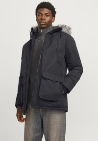 Veste noire isolée avec capuche en fourrure, dotée de plusieurs poches et d'une fermeture éclair. Associée à un col roulé sombre et à un denim.